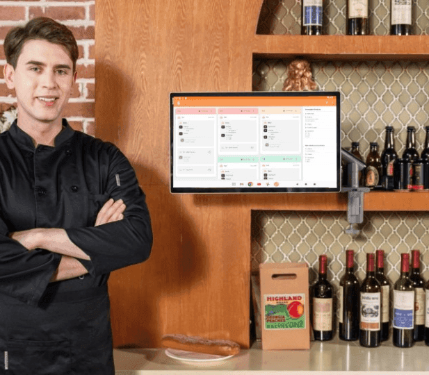 Smiling chef in black coat stands next to a monitor displaying a restaurant inventory system.